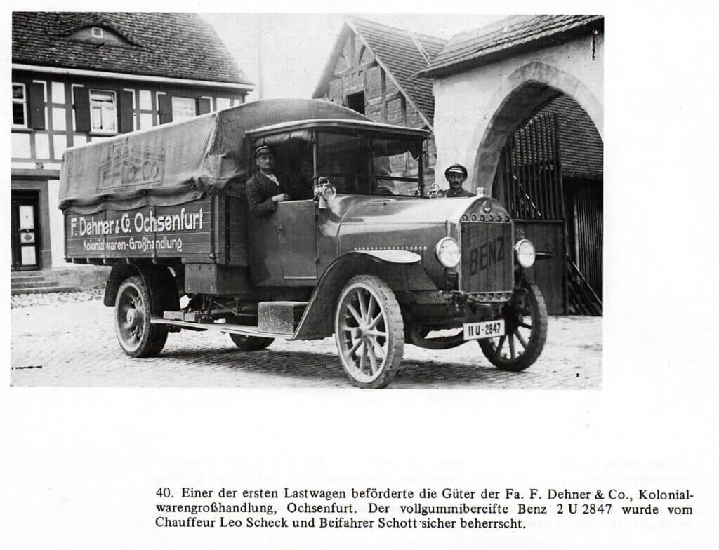 Historische Schwarzweißaufnahme eines frühen Benz-Lastwagens (Kennzeichen 2 U 2847) der Firma „F. Dehner & Co., Kolonialwarengroßhandlung, Ochsenfurt", besetzt mit Chauffeur Leo Scheck und Beifahrer Schott, vor einem Fachwerkgebäude mit Torbogen. Bildunterschrift: „Einer der ersten Lastwagen beförderte die Güter der Fa. F. Dehner & Co."