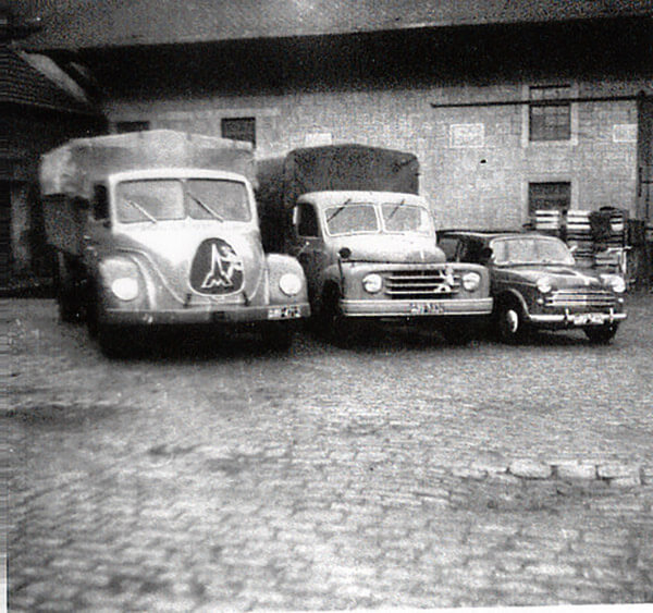 Historische Schwarzweißaufnahme von drei Nutzfahrzeugen – zwei Lastwagen mit Plane und ein Pkw – auf einem gepflasterten Hof vor einem Backsteingebäude.