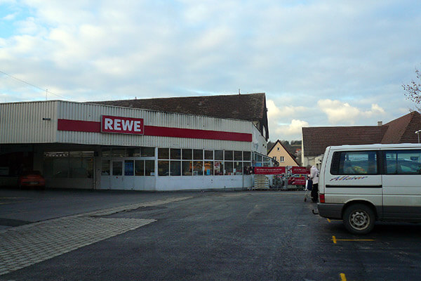 Außenansicht eines REWE-Supermarkts mit Parkplatz im Vordergrund, einem weißen VW-Bus rechts im Bild und einem Wohngebäude im Hintergrund.