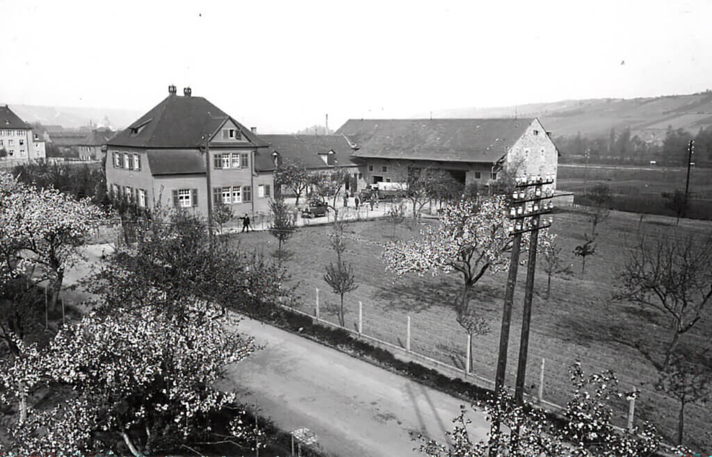 Historische Schwarzweißaufnahme eines Ortskerns mit einem zweigeschossigen Wohnhaus, angrenzenden Wirtschaftsgebäuden und blühenden Obstbäumen im Vordergrund. Im Hintergrund Hügel und weitere Bebauung.